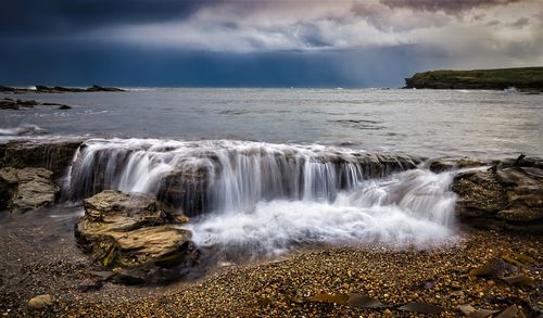 Free Scenic coastal waterfall cascading over rocks into the sea under dramatic skies. Stock Photo