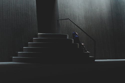 Side view of unrecognizable person sitting on concrete staircase with railing near wall of building on street with dim light