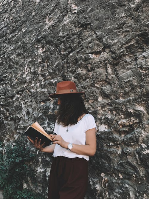 Anonymous woman reading book near stone wall