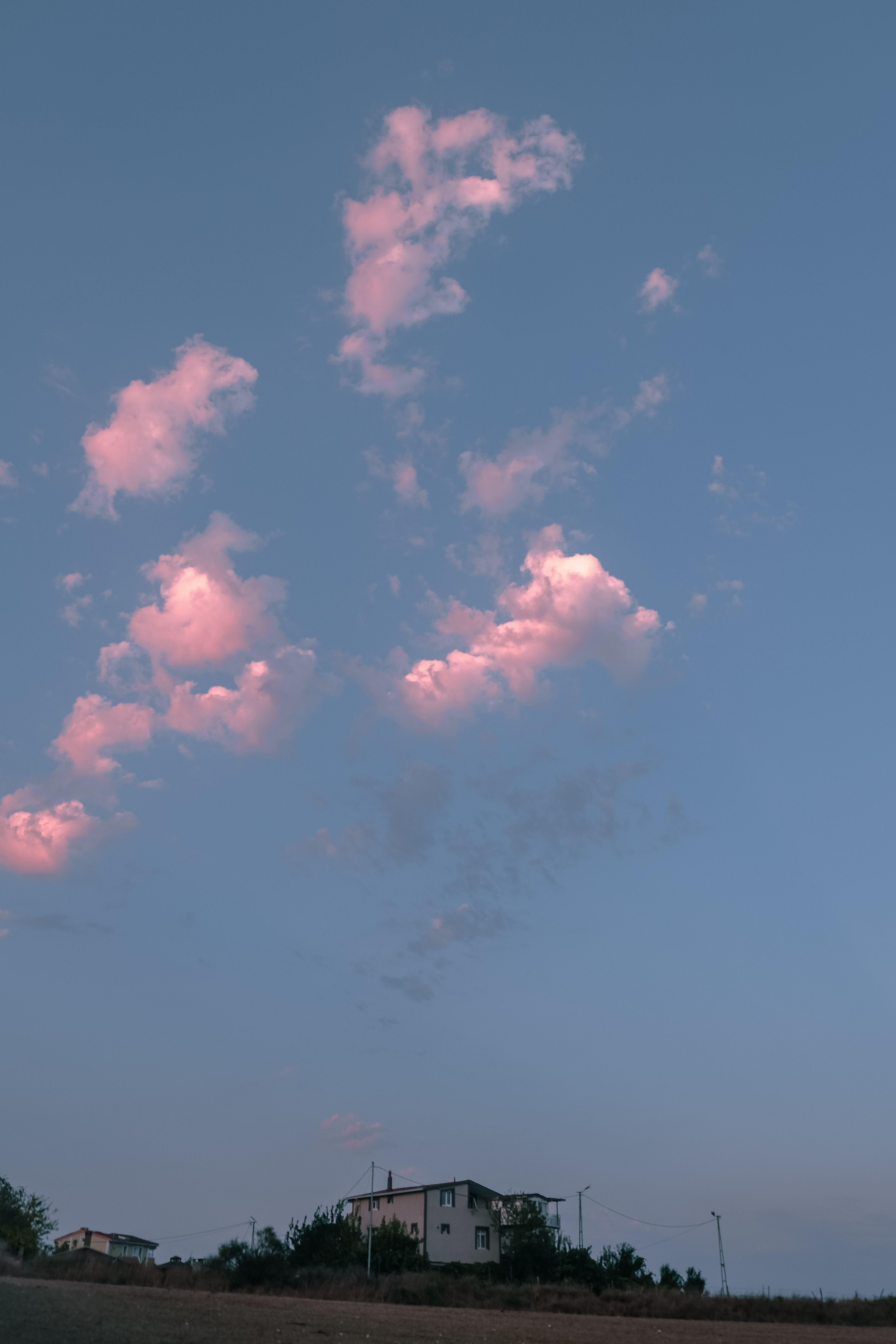 Blue sky with white clouds floating above white cottage with green trees in rural area at sundown time in countryside