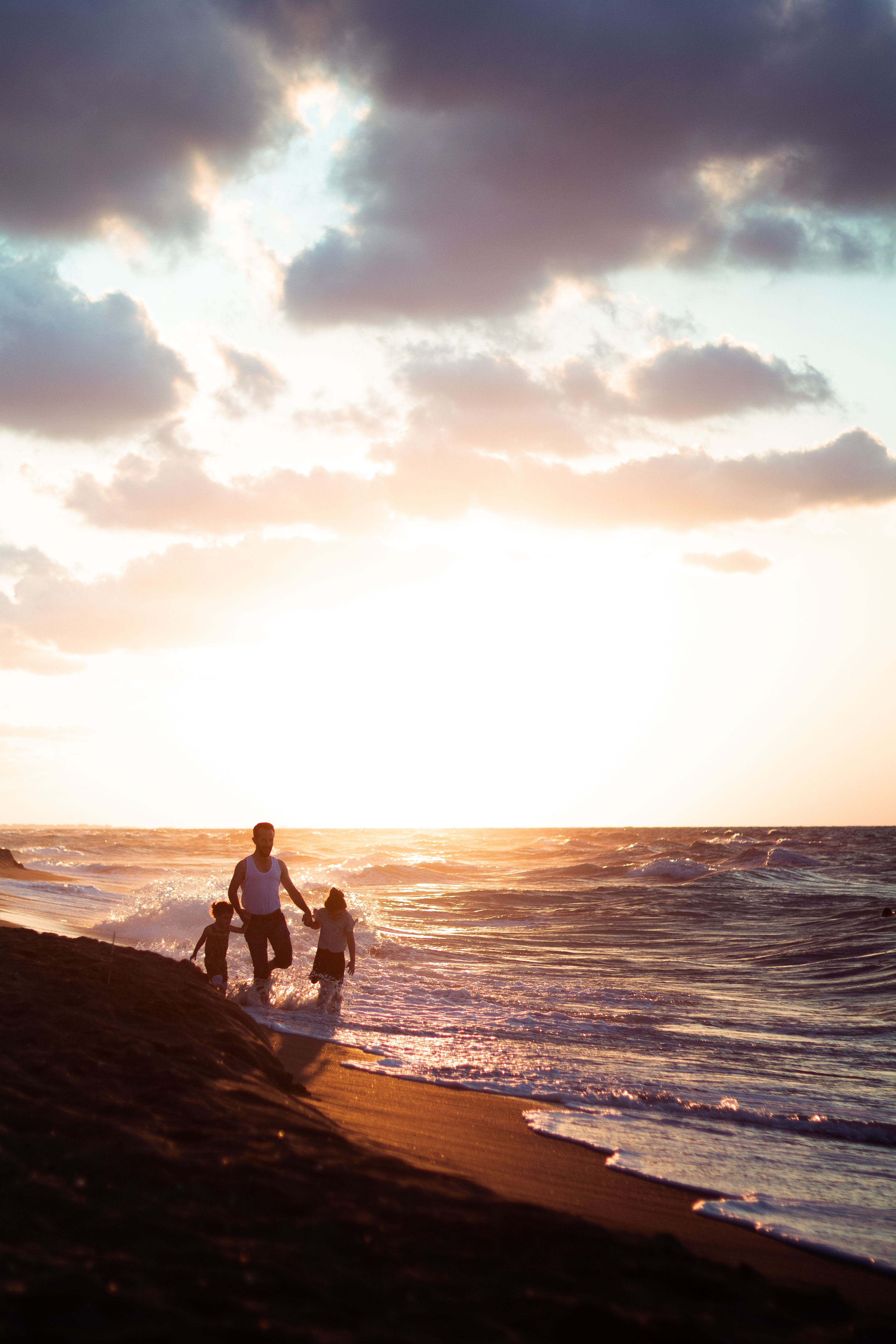 Distant father with children holding hands while strolling on sandy coast near waving sea at sundown time on summer evening