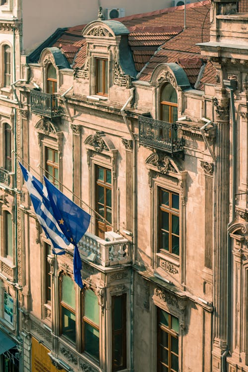 Waving flags of Greece and European union on shabby embassy building with balcony and windows on sunny street in city