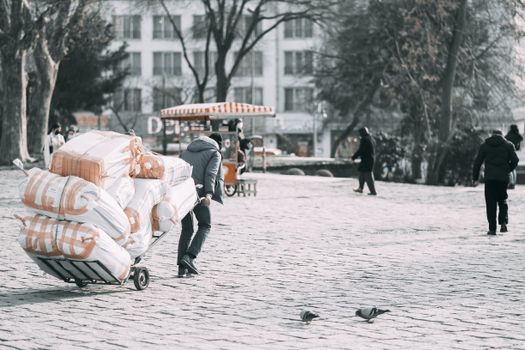 A man pulls a heavily loaded cart in a bustling İstanbul square, capturing urban life in winter.
