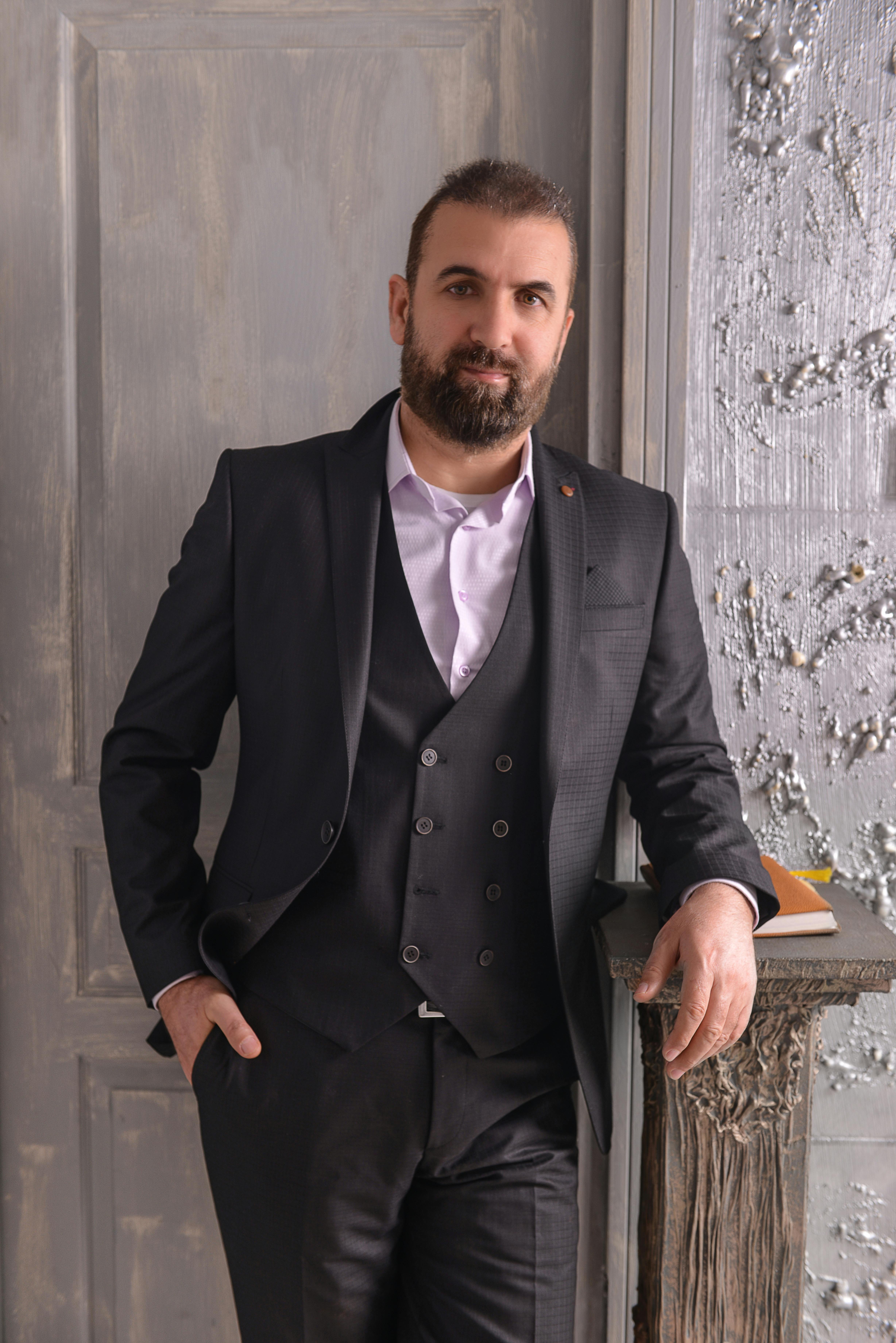 Bearded man in a stylish black suit leaning against a textured wall indoors.