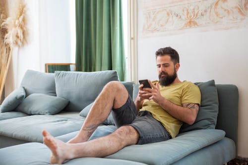 Free A Man in Yellow Crew Neck T-shirt and Denim Shorts Sitting on Sofa Stock Photo