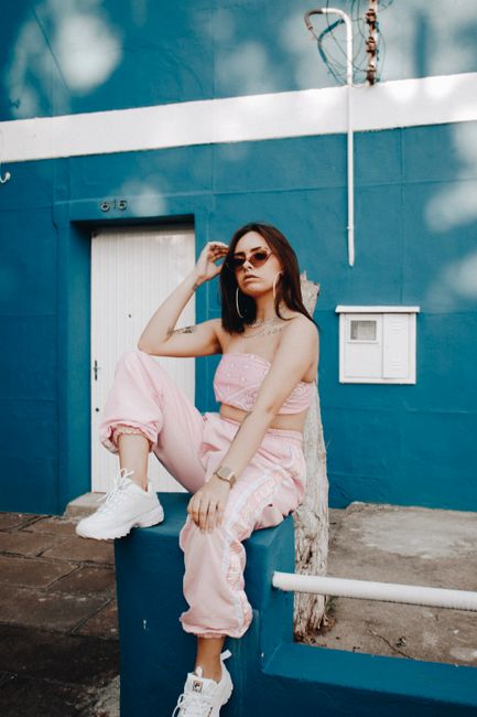 Young female in stylish outfit and sunglasses sitting on fence near blue colored wall and looking camera