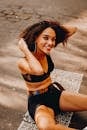 High angle of smiling ethic female in activewear sitting on asphalt road and looking at camera while resting