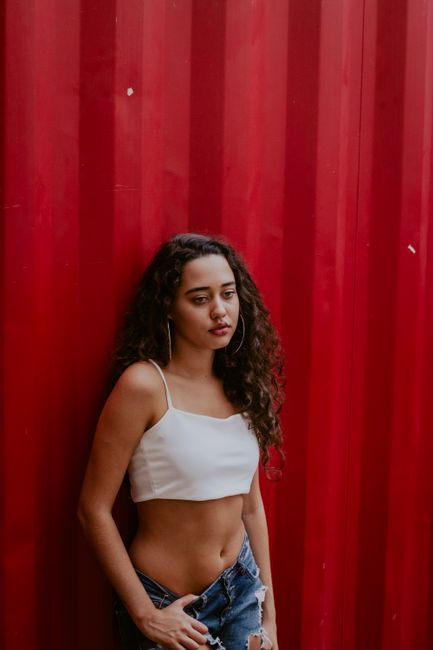 Young ethnic female with curly hair in casual clothes standing near red wall and looking away