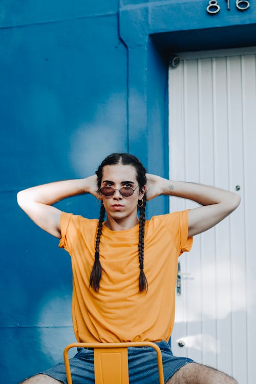 Young trendy transgender male in casual clothes and sunglasses with pigtails sitting on chair near shabby wall on street