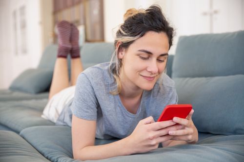 Free Smiling young woman using a smartphone while lying on a sofa at home, enjoying leisure time. Stock Photo