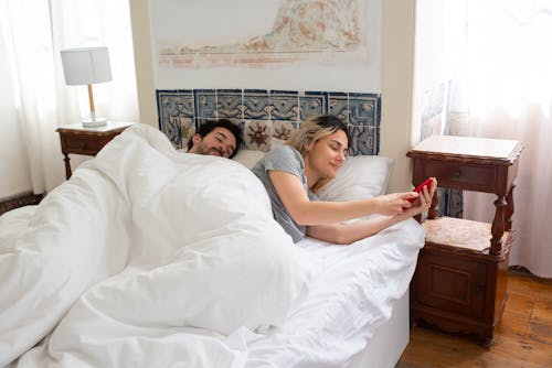 Free Woman in Gray Shirt Holding Red Mobile Phone while Lying on the Bed Stock Photo