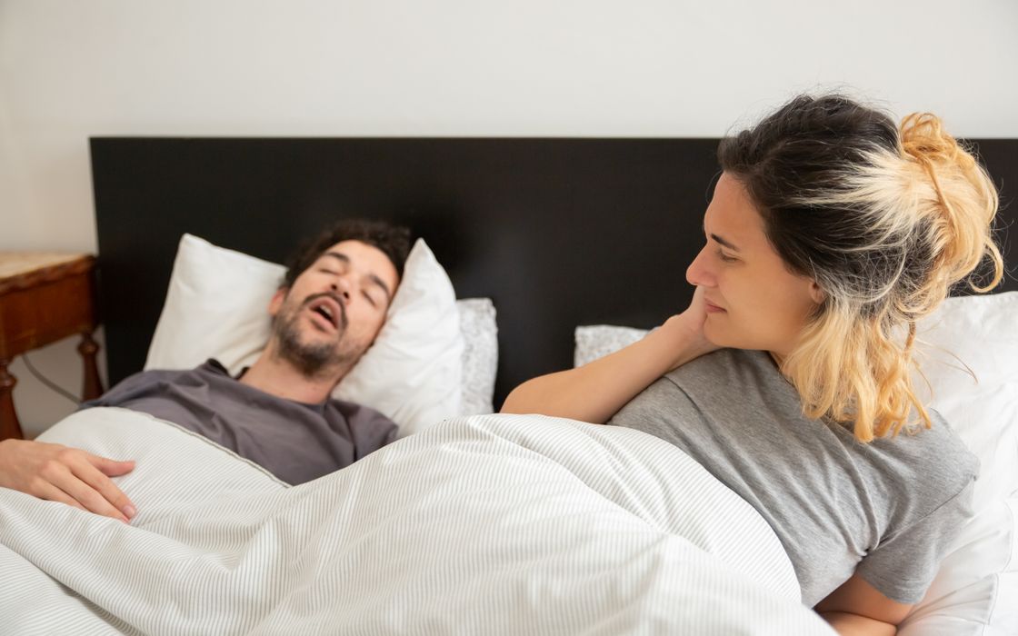 A man snores in bed while his partner lies awake beside him.