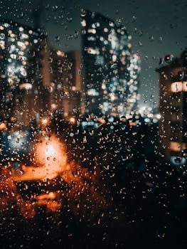 Dramatic view of a rainy cityscape at night through a wet window with blurred lights.