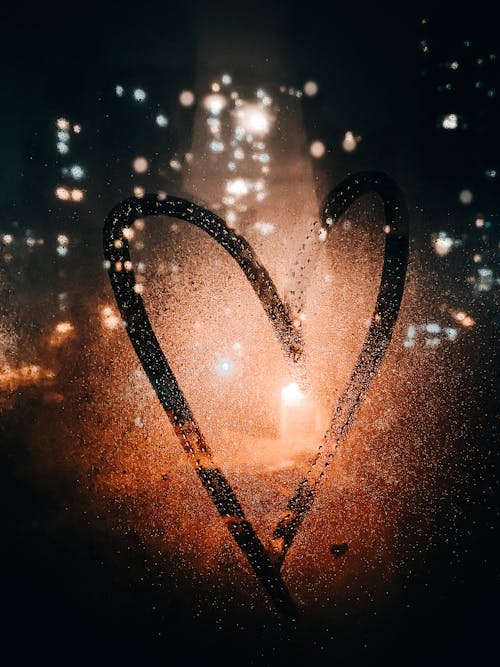 Heart drawn on glass of wet window with view of light of buildings on city street at night