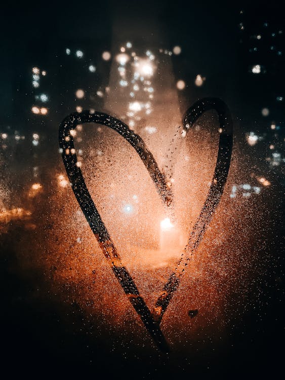 Heart drawn on glass of wet window with view of light of buildings on city street at night