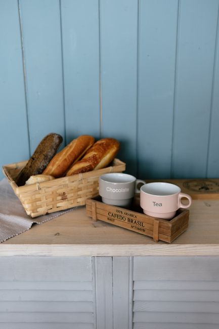 Cozy breakfast setup with a basket of fresh bread and mugs for chocolate and tea on a wooden counter.