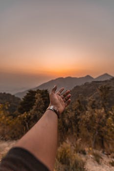 A hand reaches out towards a warm, golden sunset over the scenic mountains of Mussoorie, India.
