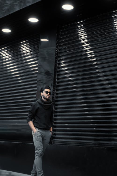 Serious bearded male in sunglasses wearing trendy clothes strolling on street with hands in pockets along building with roller shutters in city