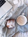 From above of cup of fresh cappuccino with sweet dessert with powdered sugar placed near laptop and TWS earbuds in case