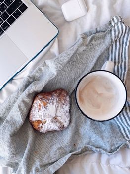 From above of cup of fresh cappuccino with sweet dessert with powdered sugar placed near laptop and TWS earbuds in case