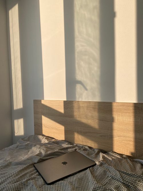 Free A cozy bedroom with soft sunlight casting shadows and a laptop on the bed. Stock Photo