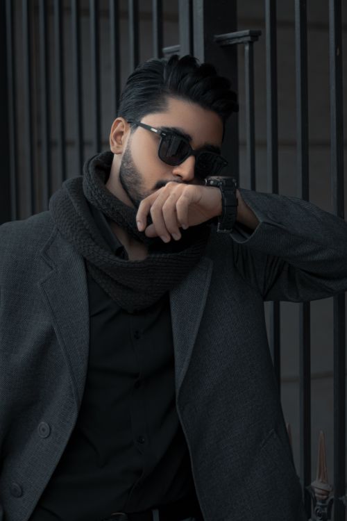 Free Stylish man in a coat and scarf wearing sunglasses, striking a pose against a wrought iron fence. Stock Photo