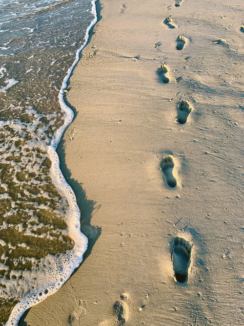 Free Footprints leading along a sandy beach as waves wash the shore, creating a serene coastal scene. Stock Photo