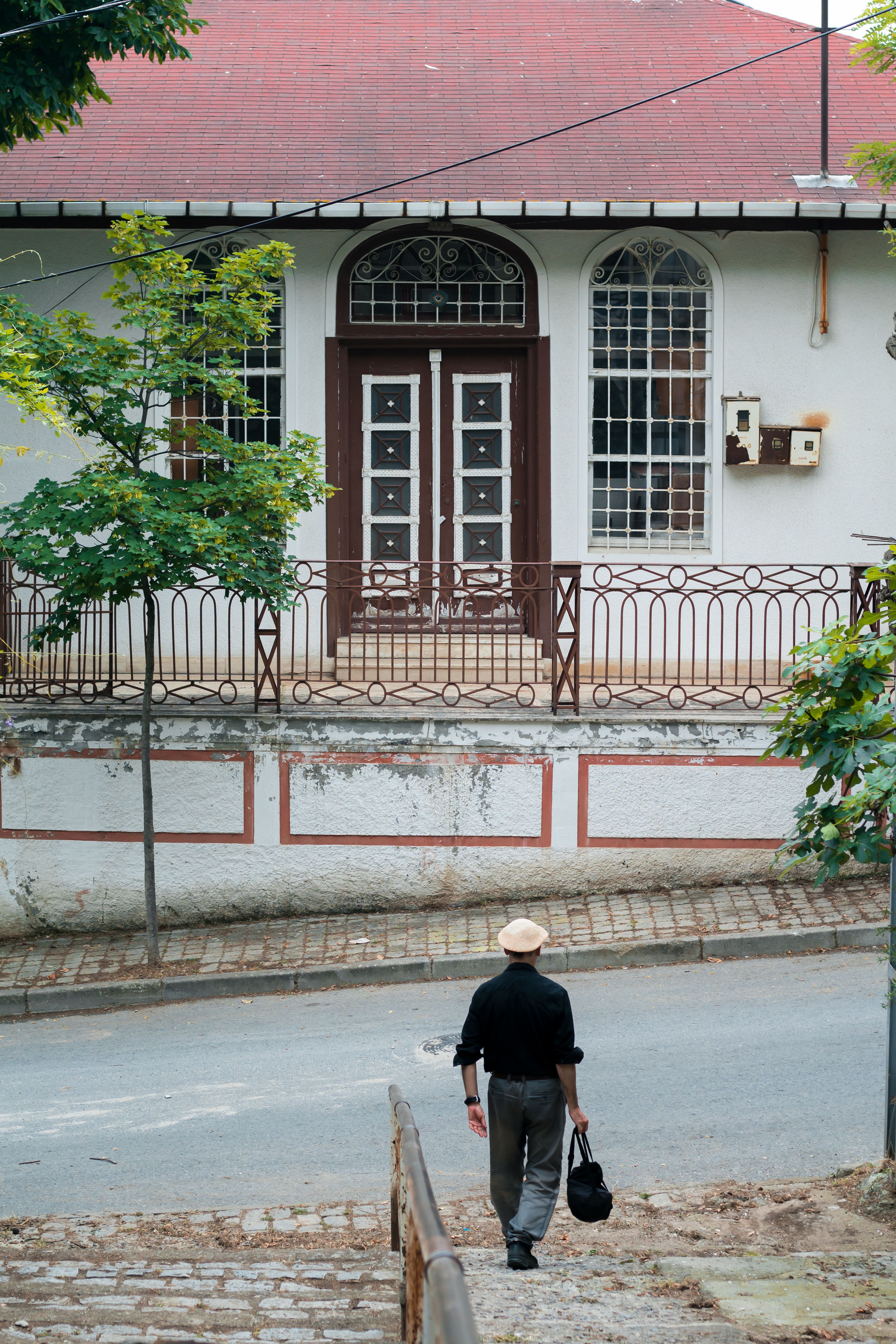 Back view of distant anonymous male in hat strolling on street with asphalt road and residential house with metal fence