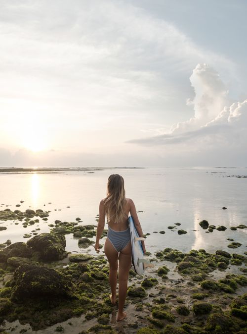 Free A woman walks towards the ocean holding a surfboard, surrounded by mossy rocks under a clear sky. Stock Photo
