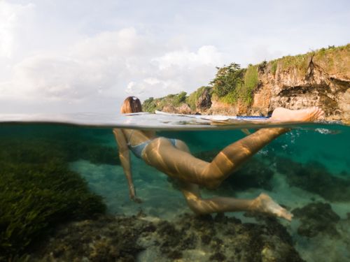 Free A woman swims underwater in a vibrant blue ocean near a rocky coastline, showcasing a serene coastal landscape. Stock Photo