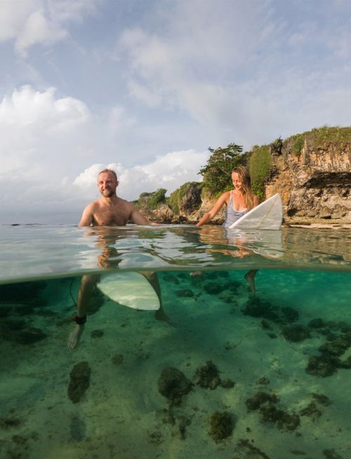 Free Man and woman with surfboards in clear turquoise waters by rocky shore. Stock Photo