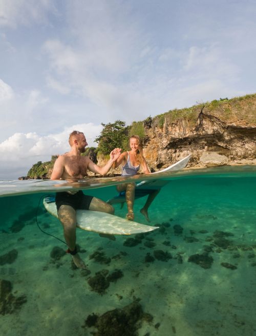 Free A couple holding hands and sitting on surfboards in crystal clear tropical waters by a rocky cliff. Stock Photo