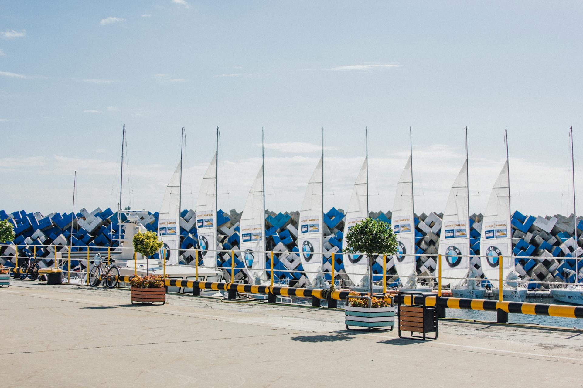 A line of sailboats docked along a waterfront with bicycles and potted plants.