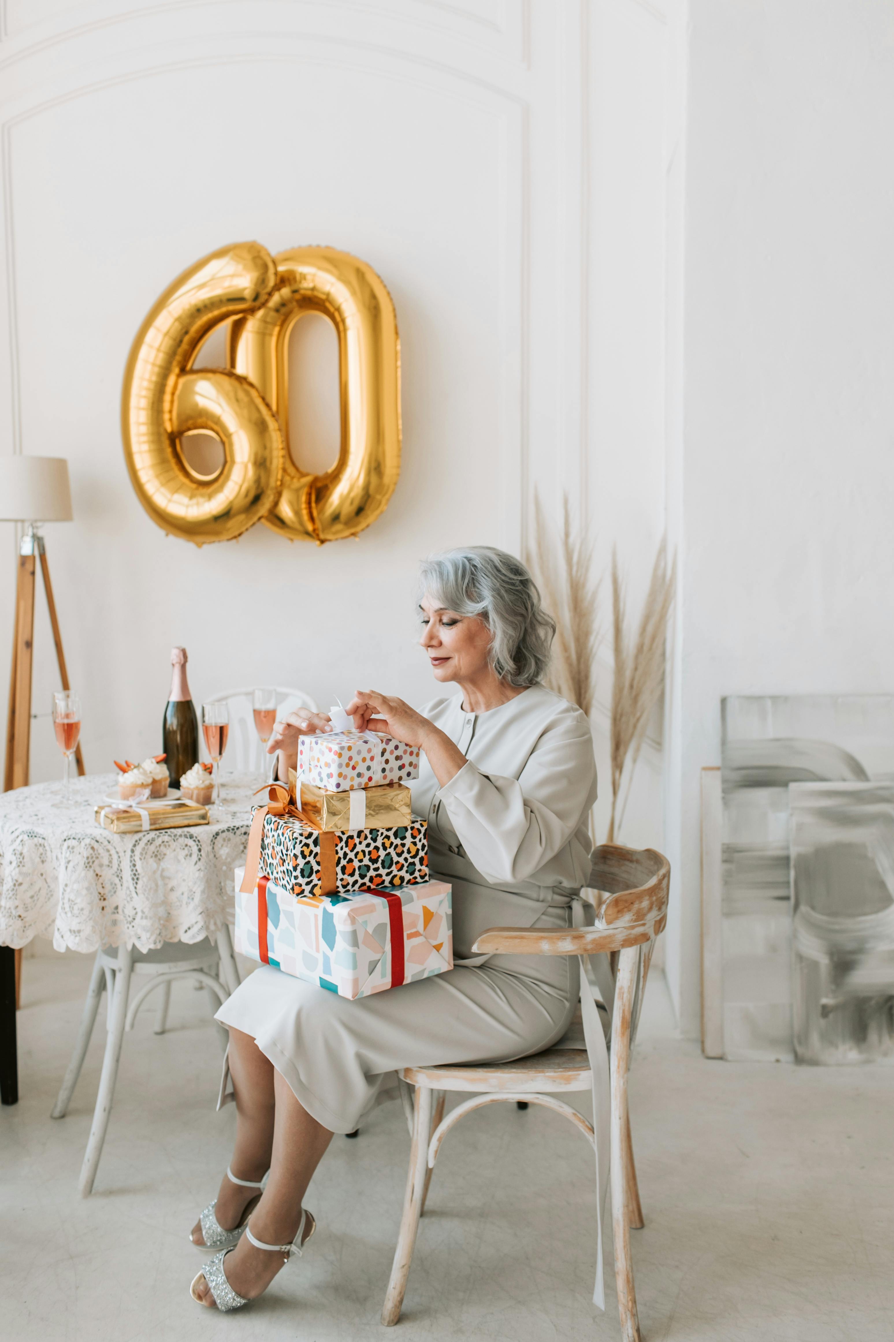 Senior woman celebrates 60th birthday indoors with gifts and champagne in a warmly decorated room.