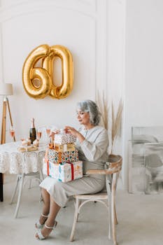 Senior woman celebrates 60th birthday indoors with gifts and champagne in a warmly decorated room.