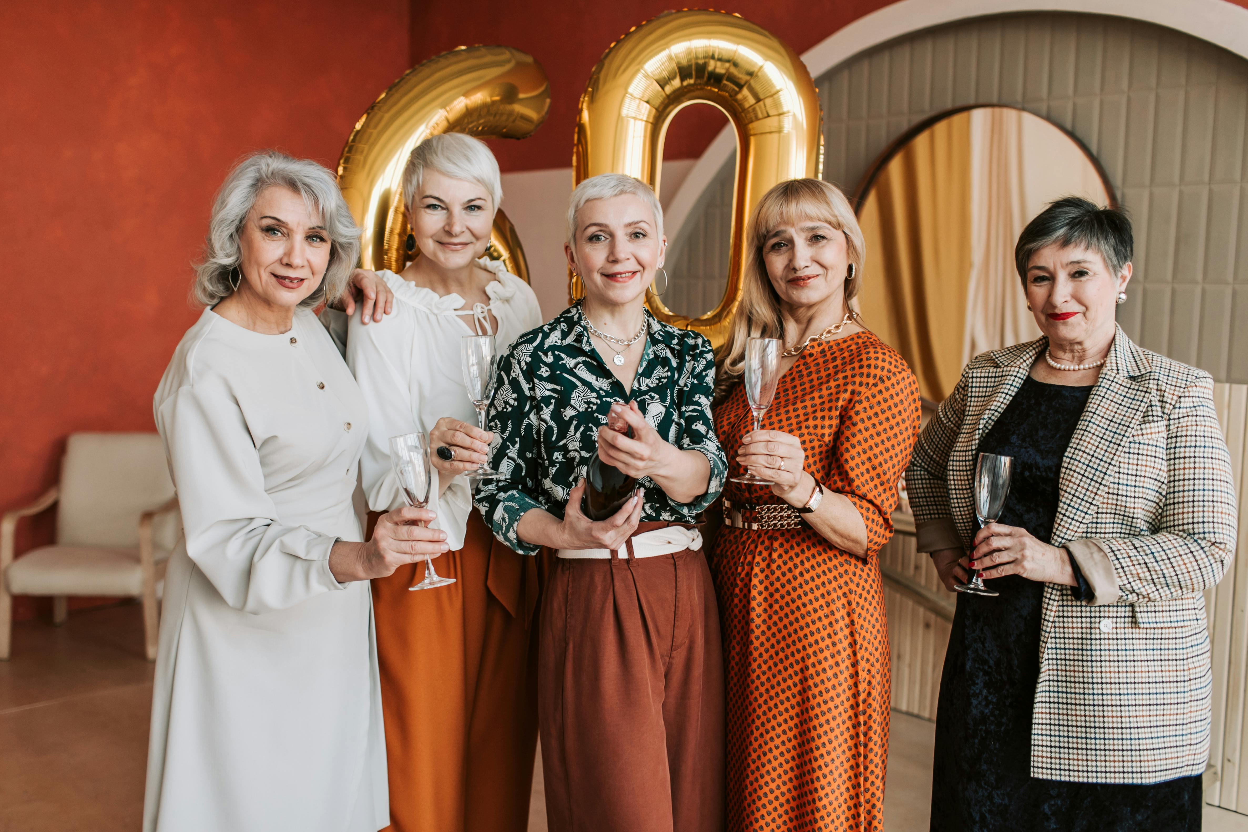 Five senior women joyfully celebrating a 60th birthday with champagne indoors.