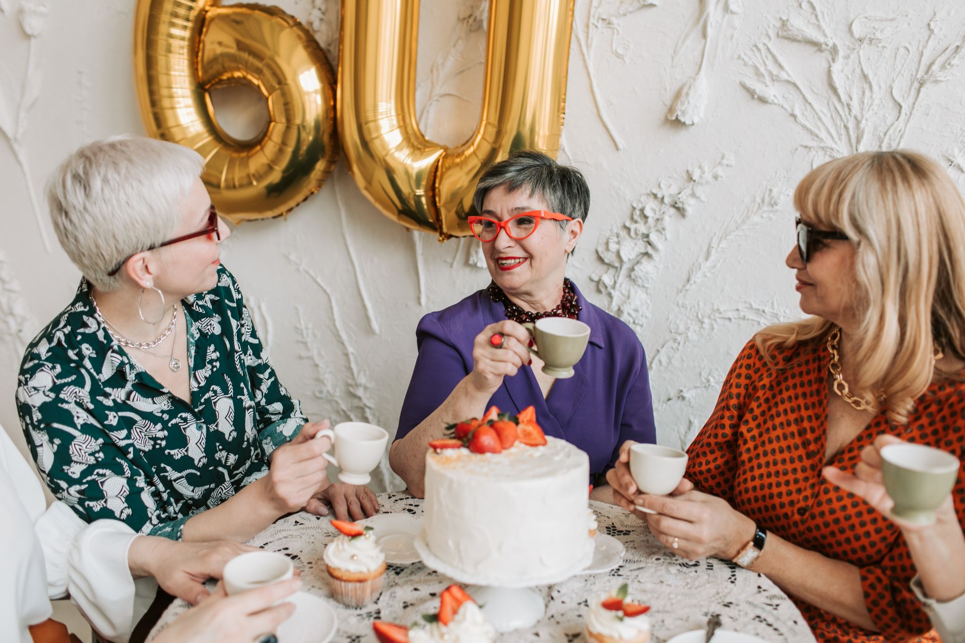Three women celebrating a 60th birthday with cake, tea, and laughter indoors.