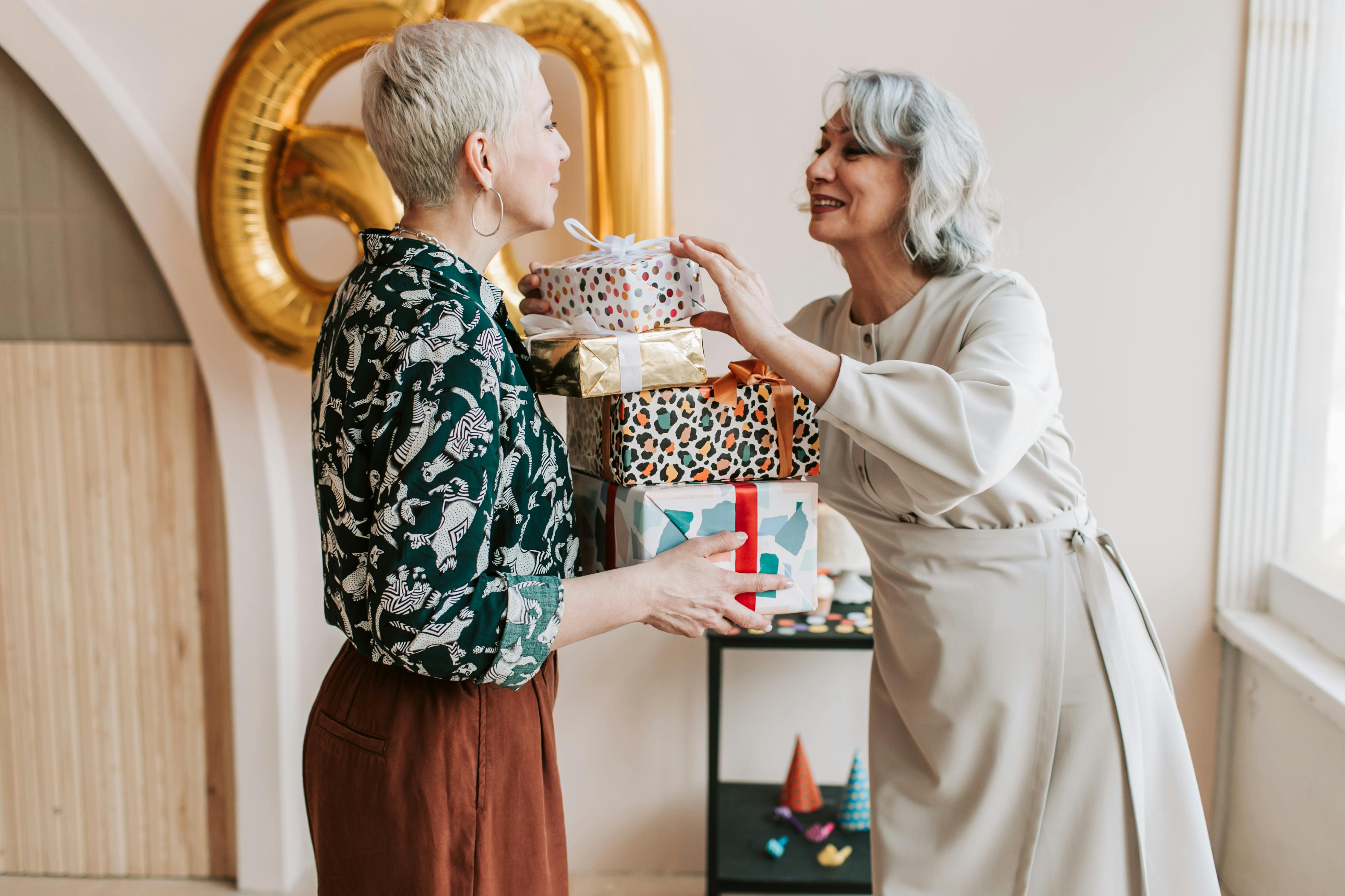 Two joyful senior women exchanging gifts during a 60th birthday celebration indoors.