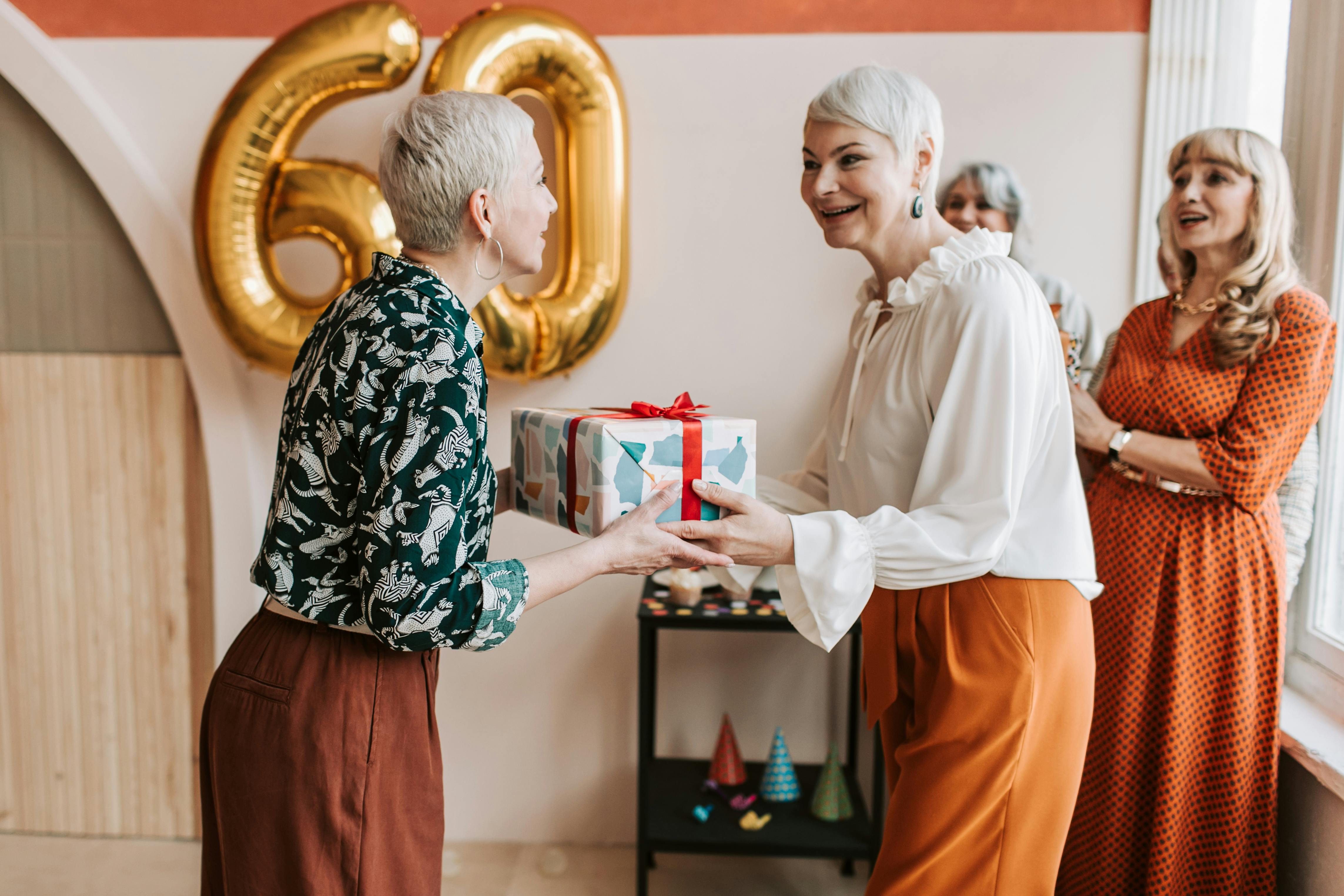 A joyful 60th birthday celebration of senior women exchanging gifts indoors.