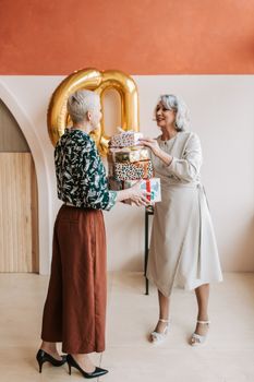Two senior women joyfully exchanging gifts at a 60th birthday celebration indoors.