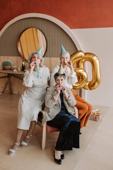 Three senior women in party hats celebrate a 60th birthday indoors with balloons and streamers.