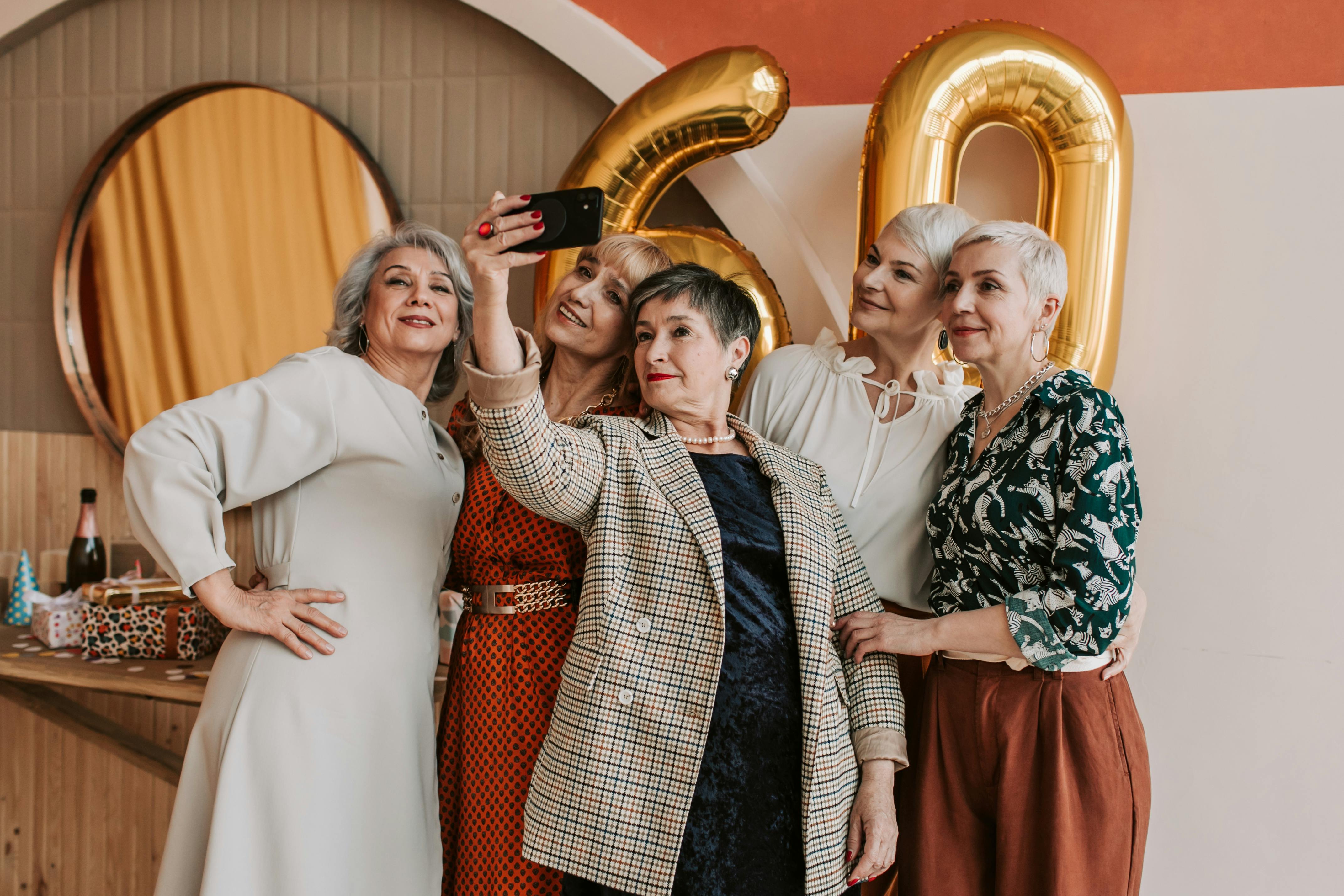 Senior women taking a selfie during an indoor birthday celebration with gold balloons.