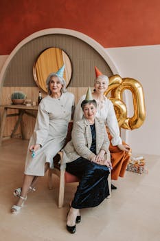 Three senior caucasian women celebrating a birthday indoors, wearing party hats and cheerful expressions.