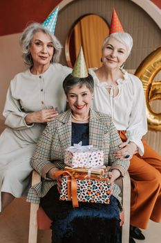 Three senior women wearing party hats, celebrating a birthday with gifts indoors.