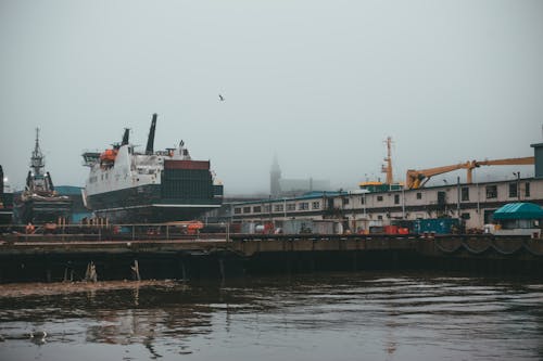 Different vessels and cranes placed on pier near rippling river in coastal area on misty day with white thick fog