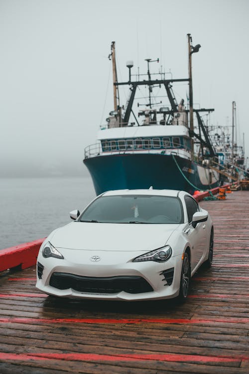 White modern automobile parked on wooden pier near moored big ship on rippling water in coastal area in foggy weather