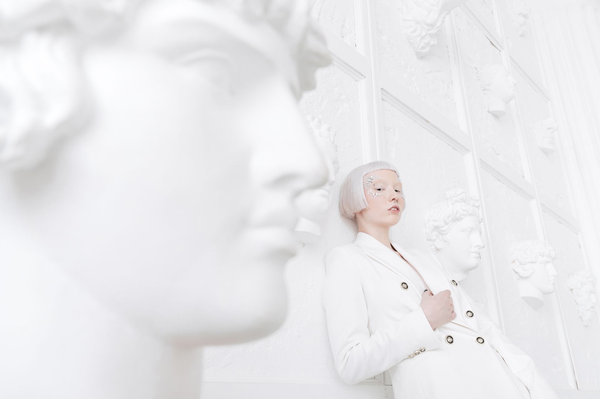 Artistic portrait of a woman with face jewels in a white suit, surrounded by classical bust sculptures.