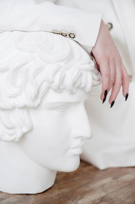 Close-up of a hand with manicured nails resting on a white sculpted bust in an artistic setting.