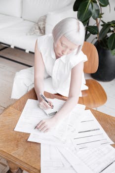 A woman with white hair sits and writes on documents at a wooden table in a bright room.