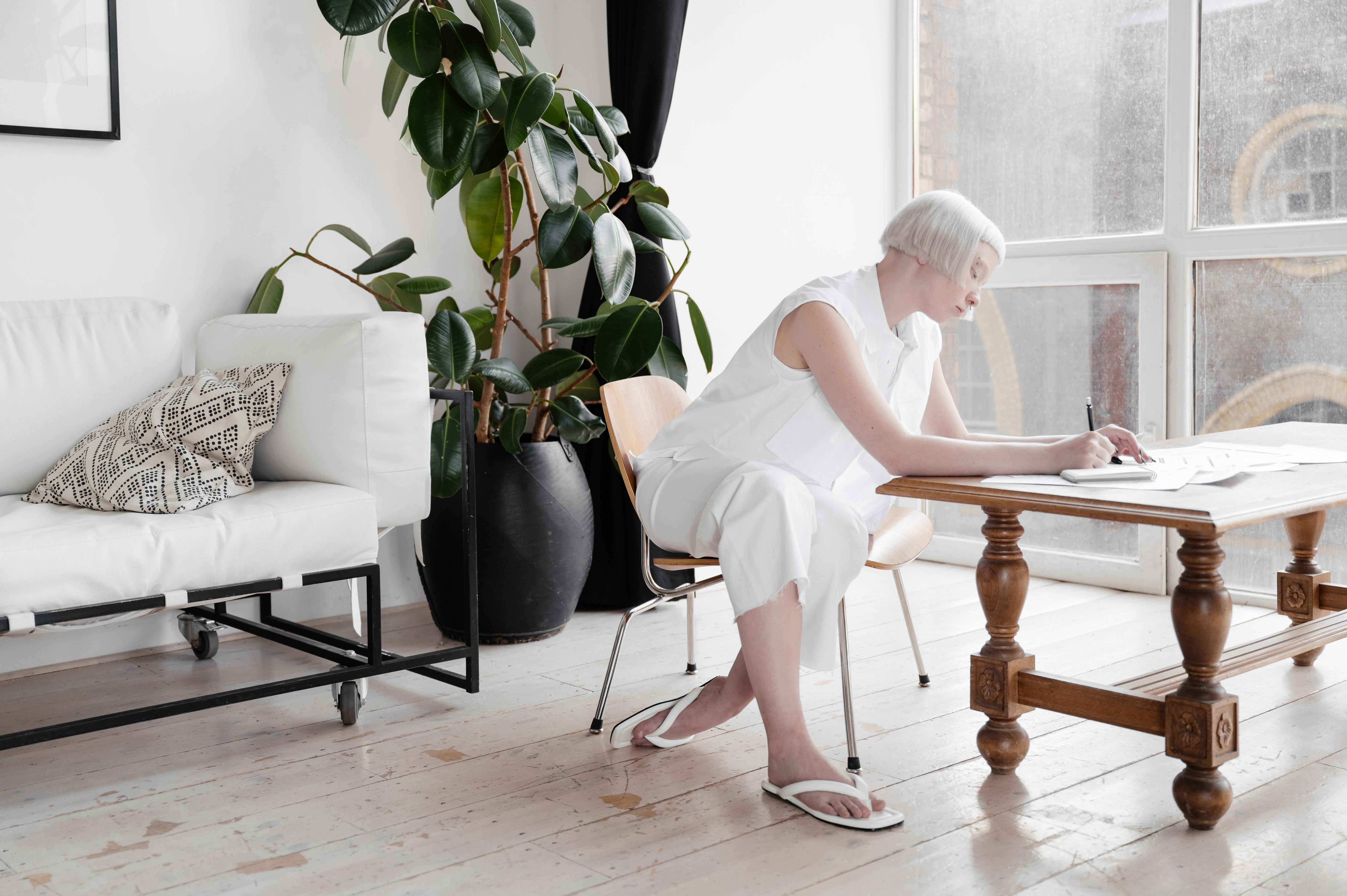 A woman with white hair writes in a bright, modern living room with large windows.